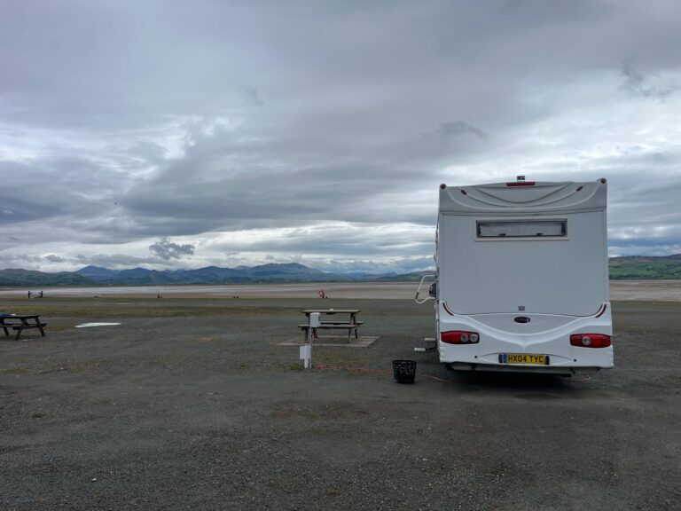 Motorhome connected to an electric hook-up on a coastal touring site with mountain views and cloudy skies.