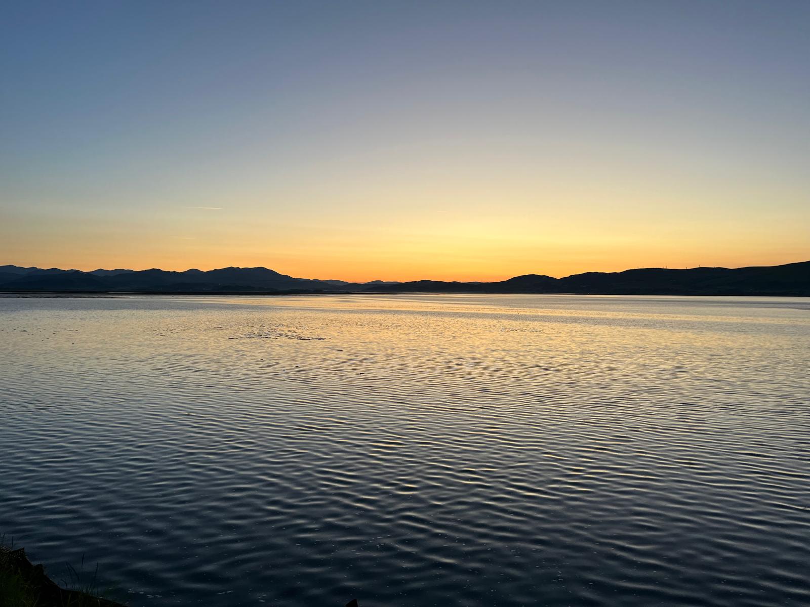 Calm coastal water at sunset with mountain silhouettes and golden evening skies reflected across the sea.