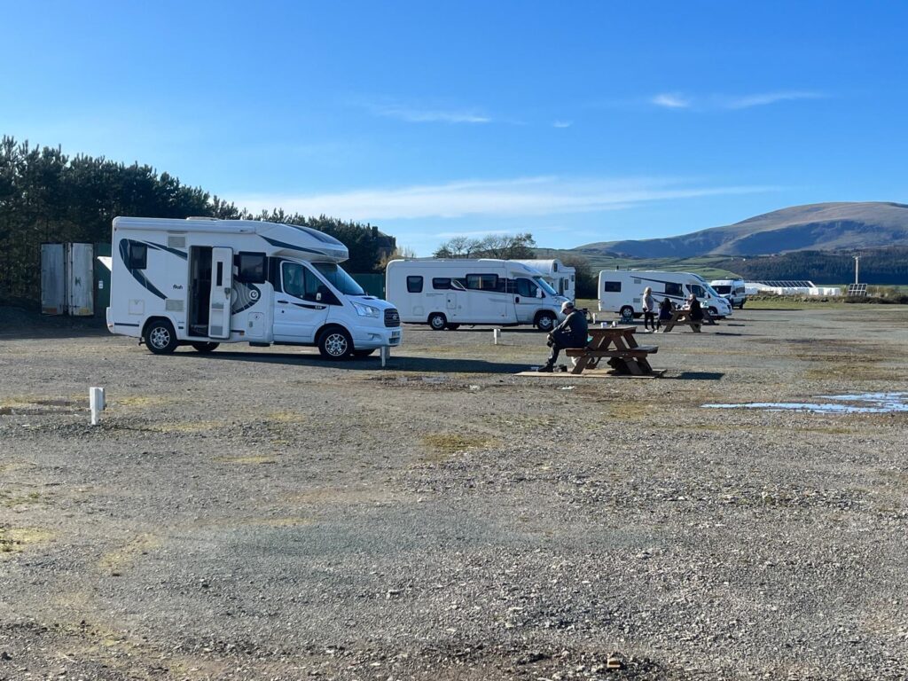Motorhomes parked on a gravel touring site with picnic benches, countryside views and clear blue skies.