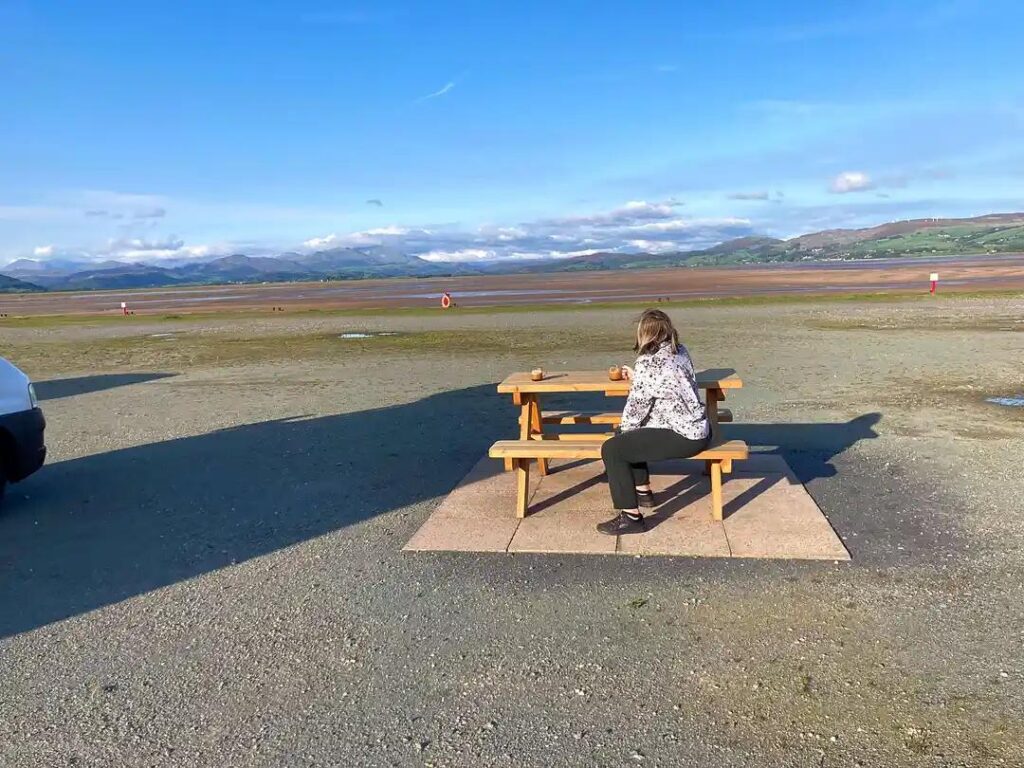 A person sitting at a picnic bench overlooking a wide coastal landscape with mountains in the distance on a bright sunny day.